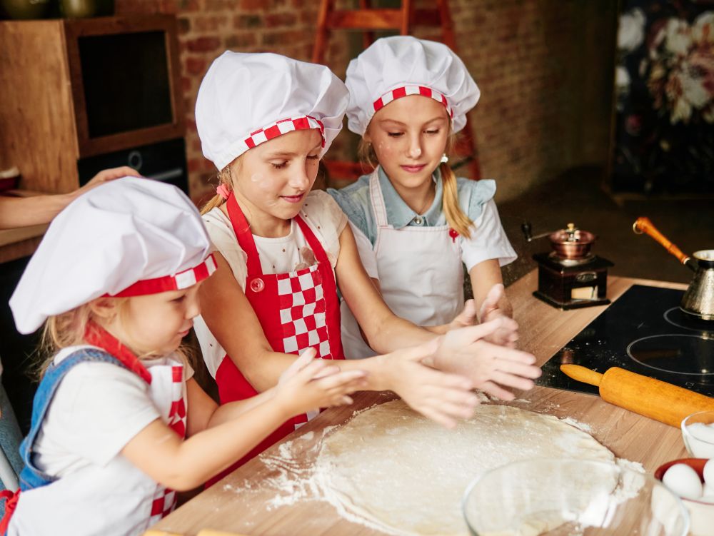 Trzy dziewczynki ubrane w fartuszki i czapki kucharskie podczas zajęć z gotowania dla dzieci w trakcie robienia ciasta na pizzę. 
