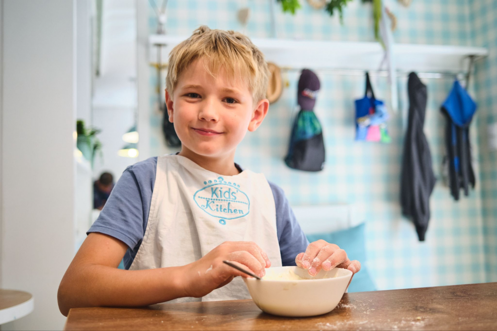 Młody, uśmiechnięty chłopiec w niebieskiej koszulce i białym fartuszku z napisem 'Kids Kitchen', trzyma miskę i miesza w niej składniki. Ręce ma obsypane mąką, a w tle widać jasną kuchnię z turkusowymi detalami.