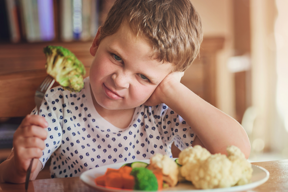 Mały chłopiec z niechętną miną przygląda się kawałkowi brokuła na widelcu, siedząc przed talerzem pełnym gotowanych warzyw.